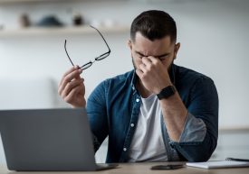 Tired young male feel pain eye strain hold glasses, rubbing dry irritated eyes, fatigued from computer work, stressed man suffer from headache, bad vision sight problem sit at home table using laptop