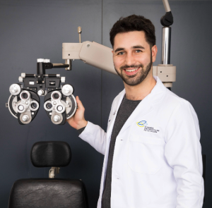 Dr. Jordan Kassouf stands in front of eye exam equipment, wearing a white lab coat over a grey professional shirt. He has black hair and neatly groomed facial hair.
